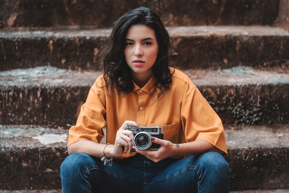 A young woman in a yellow shirt sits on outdoor steps holding a vintage camera.