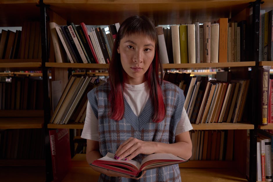 A young woman stands in a library holding an open book, surrounded by bookshelves.