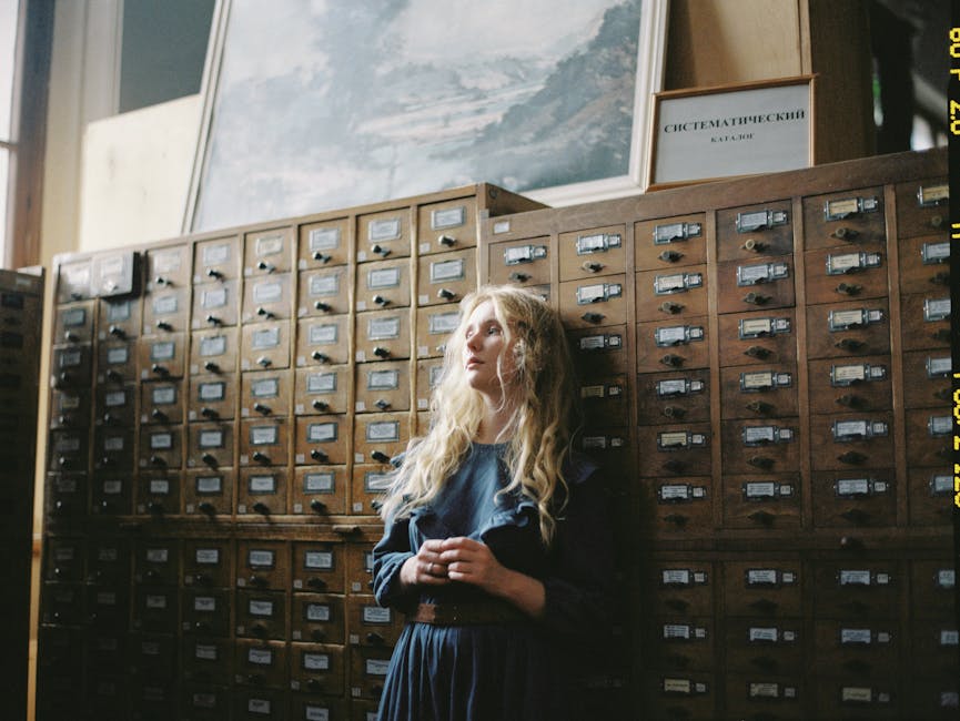 Blonde woman in a blue dress stands thoughtfully by vintage library filing cabinets.